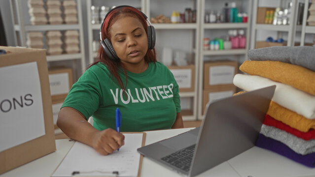 Volunteer woman wearing headphones writing on clipboard beside laptop inside a charity donation center; compassion.