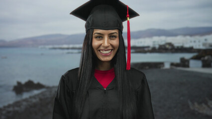 Young hispanic woman in graduation uniform gestures silence on seaside promenade with sea and beach...