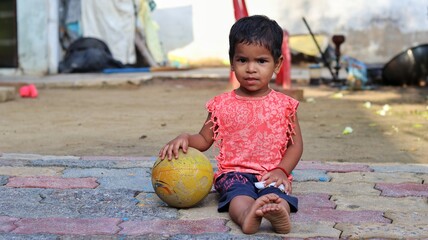 Fototapeta premium Young Indian girl sitting barefoot outdoors, holding a worn yellow ball and smiling slightly, captured in a candid moment of rural childhood