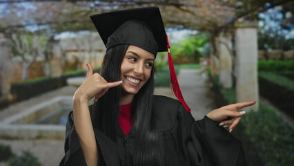 Young hispanic woman in graduation gown and cap making a phone gesture and smiling in a sunny...