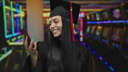 Young hispanic woman in graduation uniform holds thailand passport smiling in a casino with slot...