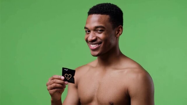 Young african american man with a bare chest smiling confidently while holding a condom on a green screen, promoting safe sex, contraception, and sexual health awareness