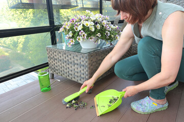 Woman rakes trash on dustpan, dry leaves and flowers on garden terrace in summer. Terrace in backyard. Cleanliness and order.