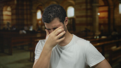Man pinching nose with eyes closed in dimly lit stone church with arched windows; discomfort...