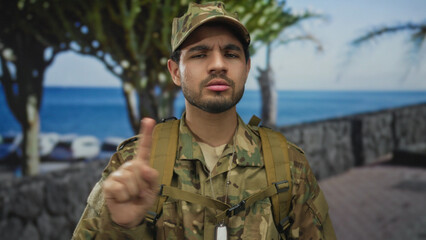 Man in camouflage uniform and cap raises index finger on street promenade under tropical palm...