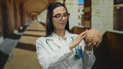 Hispanic woman doctor in uniform holding brain model with stethoscope in old university outdoor...