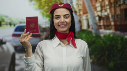 Woman holding red passport in front of a street background wearing flight attendant uniform with...