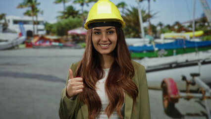 Hispanic woman wearing hardhat and suit gives thumbs up on a scenic beach while gesturing towards...