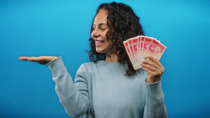 Middle-aged woman holds israeli shekels joyfully against a blue background, extending and pointing...