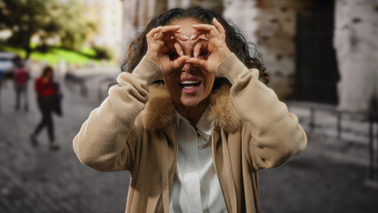 Fototapeta premium Mature hispanic woman with curly hair playfully gestures binoculars with her hands on a sunny city street.