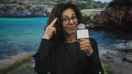 Middle aged hispanic woman reporter points finger to head holding microphone at beach; thinking...