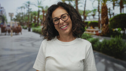 Middle aged hispanic woman with curly hair and glasses opens mouth in playful grin on street; joy...