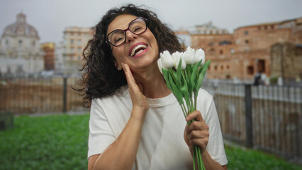 Woman holding bouquet of white flowers in front of roman ruins outdoors with joyful expression in...