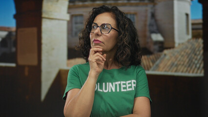 Woman wearing green volunteer shirt stands in thoughtful pose on charming old town street outdoors,...