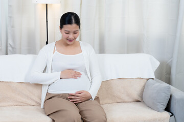 Asian mother to be sitting calm smiling warmly gently placing hands on belly wearing comfortable...