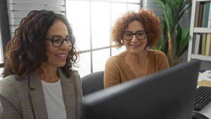 Hispanic women working together in a modern office setting, showcasing professional partnership and...