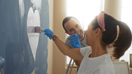 Hispanic couple decorating new home together painting wall indoors man and woman smiling creating...