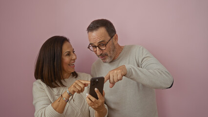 Hispanic couple in love sharing joyful moment on smartphone against isolated pink background...