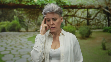 Woman touching temple with thoughtful gaze near a mossy stone path in vibrant green park;...