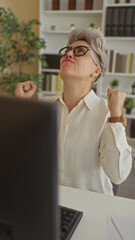 Woman in white shirt and glasses raises fists by computer desk in sunlit office surrounded by...