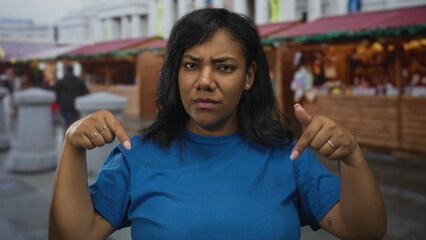 Woman pointing finger downward at her blue shirt in busy outdoor market while frowning; assertion...
