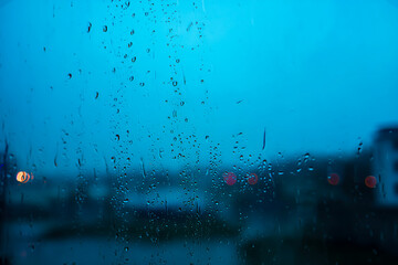 Rainy window with raindrops and vertical streaks, raindrops creating textured patterns on glass, soft city lights blurred in background, blue tone dominates calm rainy urban scenery