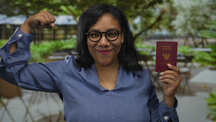 African american woman flexes arm showing bicep while holding a thailand passport amid leafy...