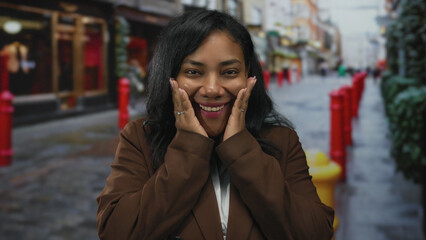 Woman pressing cheeks with hands while wearing brown coat on a cobblestone city street lined with...