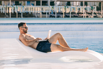 A freelancer works online on a laptop while lying on a sun lounger by the pool, enjoying summer....