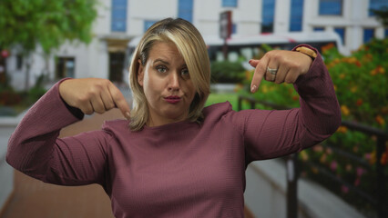 Woman with blonde hair and casual attire pointing both index fingers down at pavement on city...