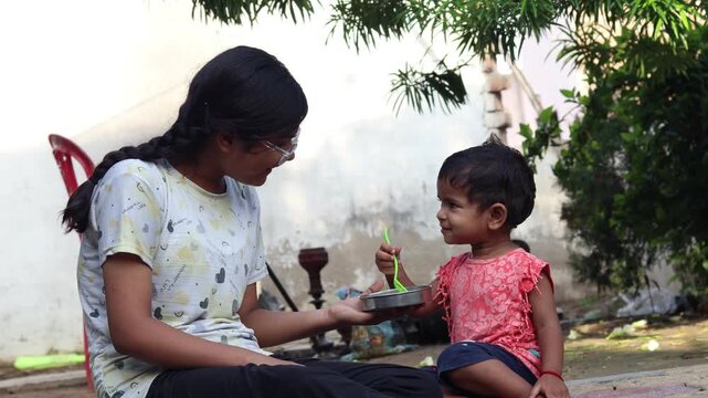 Young sisters sitting , the elder lovingly holding the younger as she eats banana from a bowl, a candid moment of sibling bonding