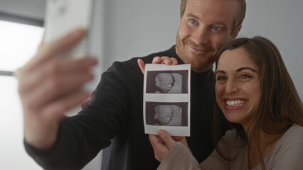 Man and woman joyously taking selfie with ultrasound photo in bright home showcasing pregnancy announcement and new family beginnings.