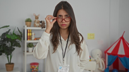 Woman teacher in preschool classroom with glasses on looking serious surrounded by toys and...