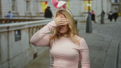 Young blonde woman in bunny ears covers her mouth in a playful moment on a city street outdoors.