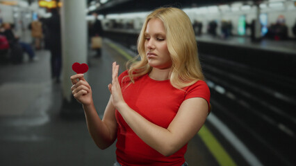 Young woman with blonde hair holds a red heart in a tube station, expressing rejection, showcasing...