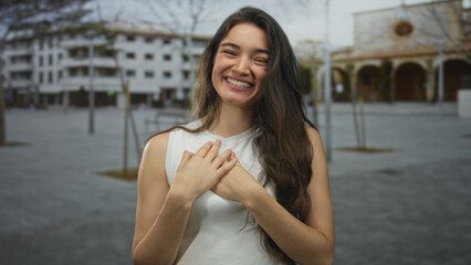 Hispanic woman touches her chest with hands and smiles on a city street in front of a building;...