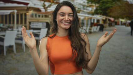 Hispanic woman smiling broadly and raising her hands with open palms at outdoor restaurant terrace;...