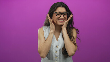 Young hispanic woman holding cheeks with clasped hands and grimacing in a purple studio for...