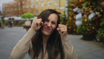 Young hispanic woman rubbing fists under eyes for crying gesture on street plaza; mockery sadness...