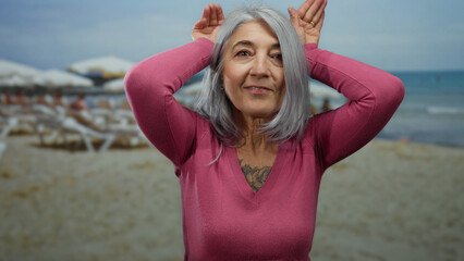 Senior woman with grey hair in pink sweater playfully makes ear gestures on a lively beach setting...