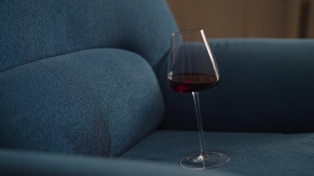 Glass of red wine falling on couch. Close-up of spilling red wine from glass onto blue couch creating large, unsightly stain, moment of carelessness and frustration of unfortunate mishap indoors.