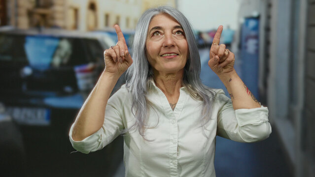Senior woman with grey hair outdoors on a street joyfully raising her fingers as if pointing at something inspiring above the city landscape.