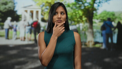 Fototapeta premium Young latin woman in a park has an idea while standing outdoors on a sunny day, surrounded by blurred people and greenery.