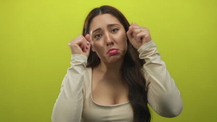 Young woman with long hair in white blouse pouts lips and makes fists by eyes in green studio; sadness.