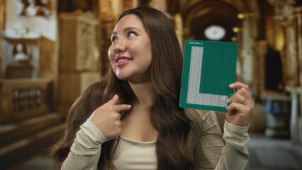 Young hispanic woman holding a green learner sign and pointing to her chest in a building;...