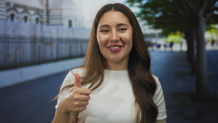 Young smiling woman showing thumb in thumbs up gesture on street lined with fence and trees;...