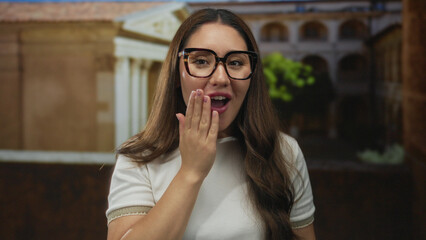 Woman wearing black glasses touches mouth with manicured hand on old town street in daylight;...