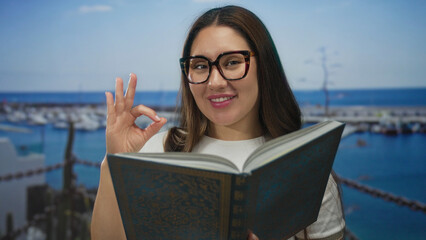 Woman reading an ornate open book with thick framed glasses on street by port boats under clear...