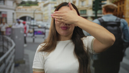 Young hispanic woman wearing glasses covers eyes with hand in front of building on busy urban street; curiosity.