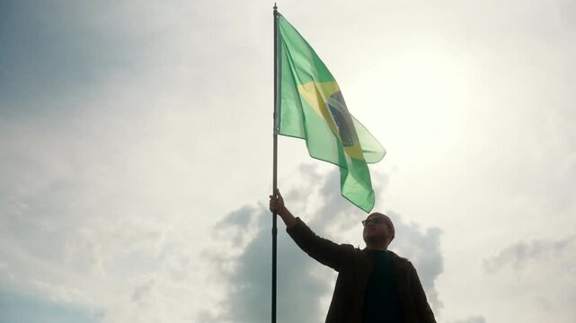 Brazilian young man standing proudly with holding and raising brazil flag against blue sky with sunlight. Proud Brazilian citizens man holding the Brazil flag waving celebration Independence Day.